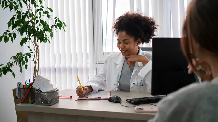 Smiling female doctor with curly hair in white coat writing notes at desk while consulting with patient in bright office with plants and window blinds